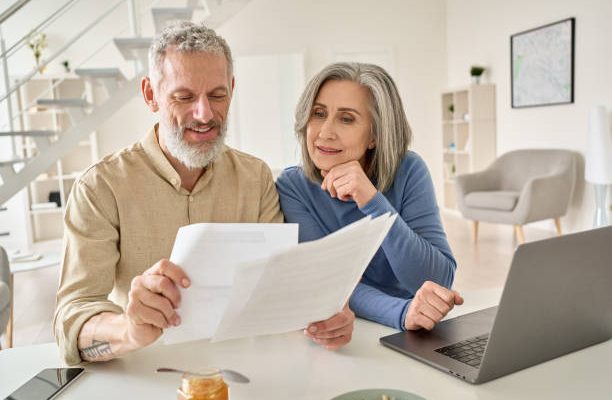 Middle aged senior old couple holding documents reading paper bills paying bank loan online, calculating pension fees, payments, taxes, planning family retirement money finances using laptop at home.